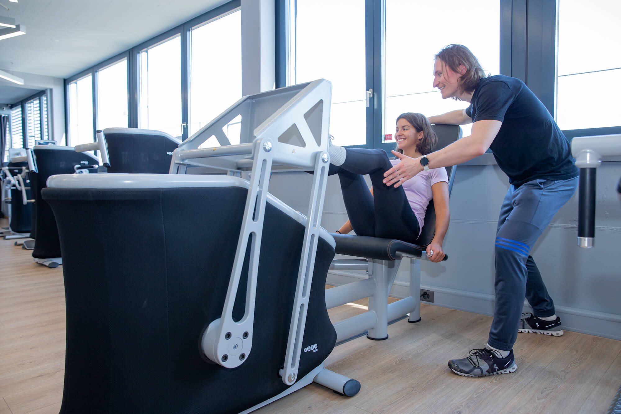 Patientin trainiert an einem Bein- und Hüftgerät, ein Physiotherapeut betreut sie in einem hellen Trainingsraum