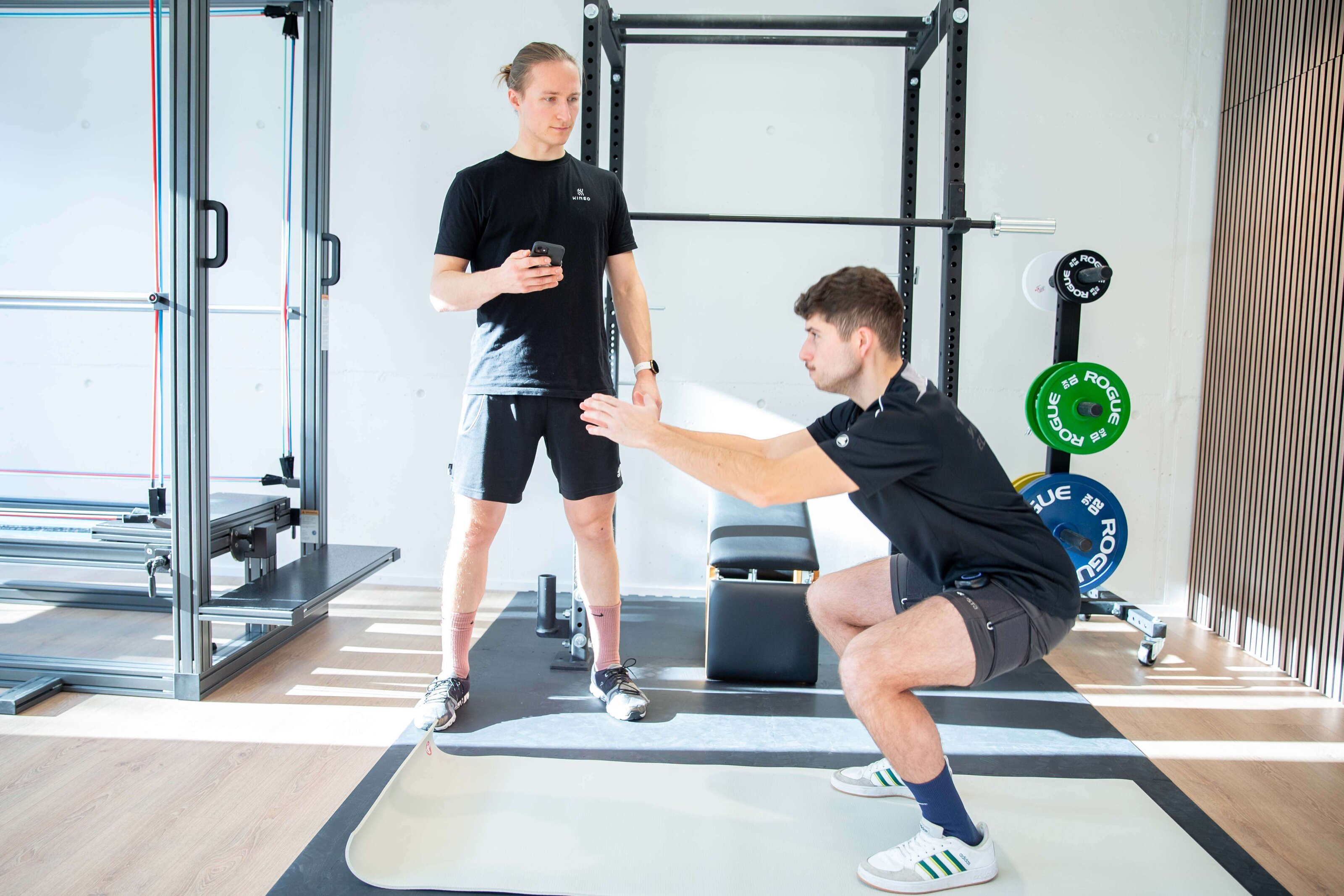 Athlet bei der Kniebeuge im Rack, ein Physiotherapeut beobachtet die Ausführung