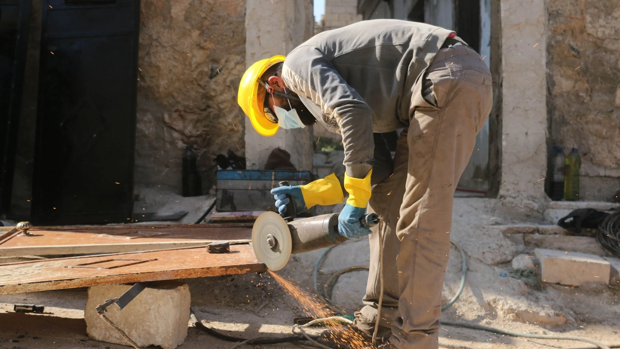 Pracownik budowlany noszący żółty twardy kapelusz, maskę na twarz i rękawiczki używające szlifierki elektrycznej na blachy, lecą iskry.