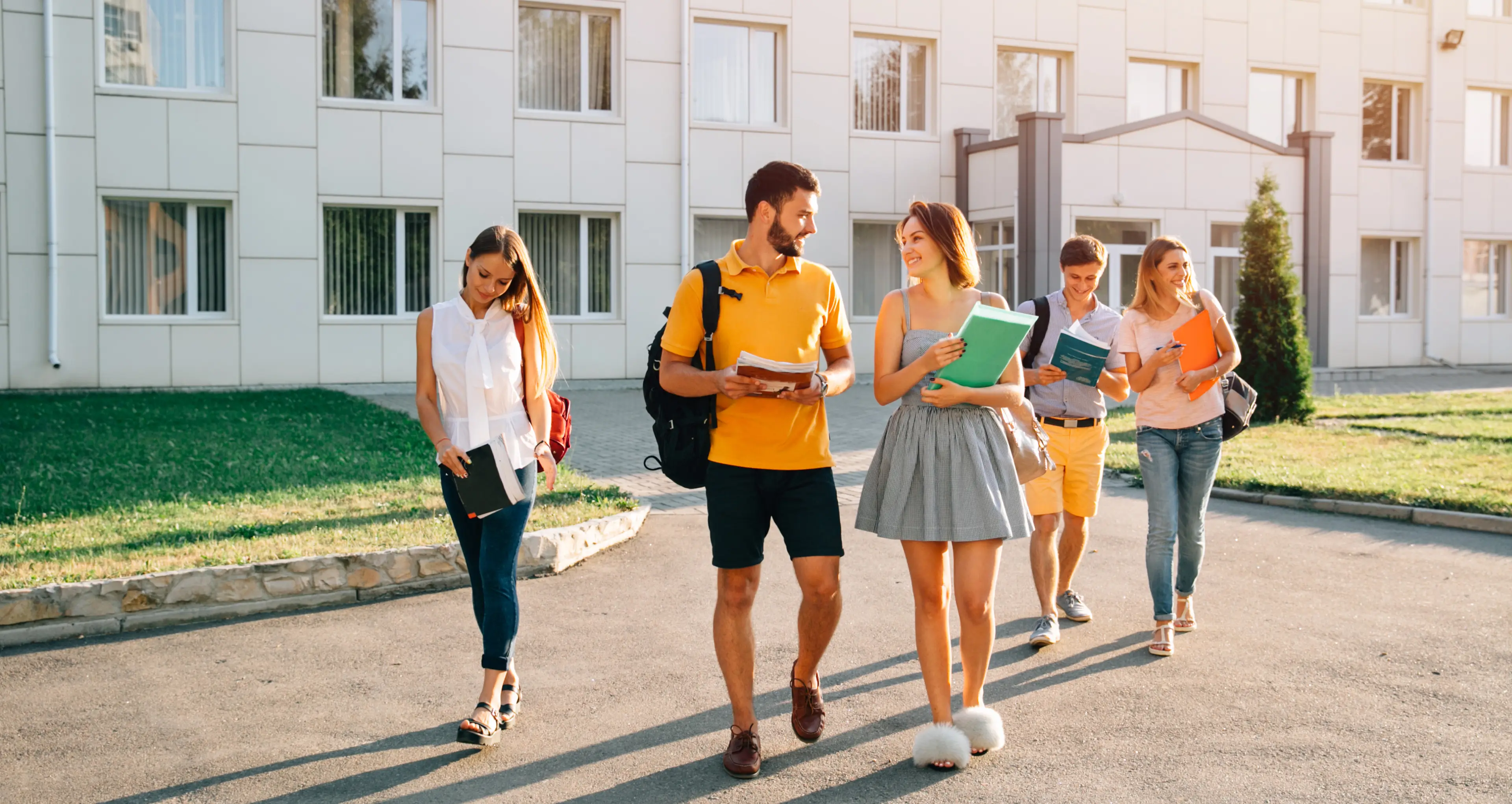 Grupa pięciu różnych studentów chodziła na miejscu w pobliżu budynku, trzymając książki i uśmiechając się.