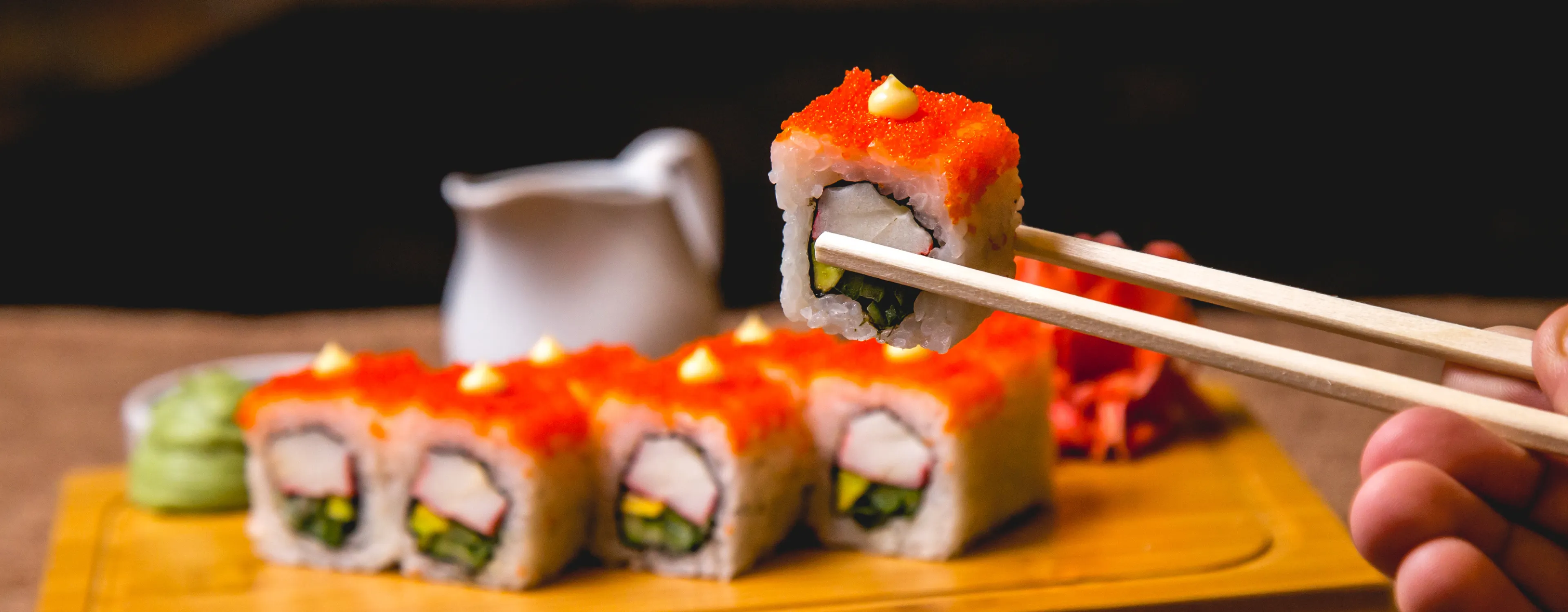 Ręczne trzymanie pałeczek chwytających bułkę sushi zwieńczoną pomarańczową ikrą rybną i małą porcją sosu, z większą ilością bułek sushi i wasabi na drewnianej desce w tle.