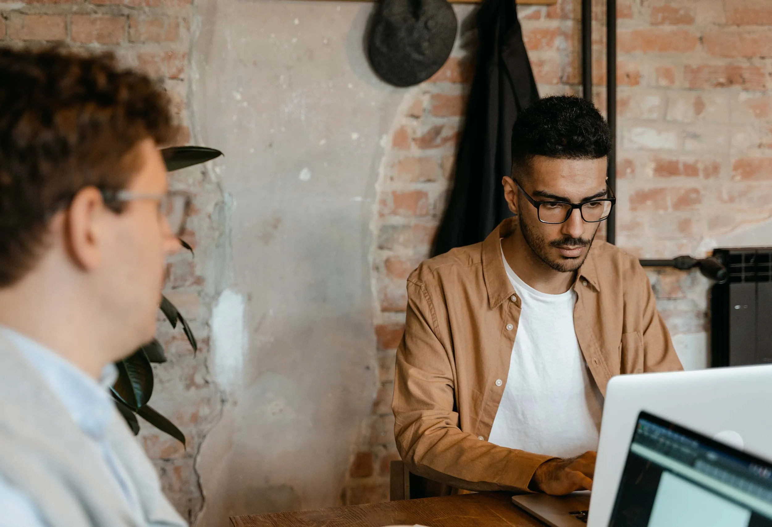 Dwóch mężczyzn pracujących na laptopach w rustykalnym biurze z odsłoniętymi ceglanymi ścianami.