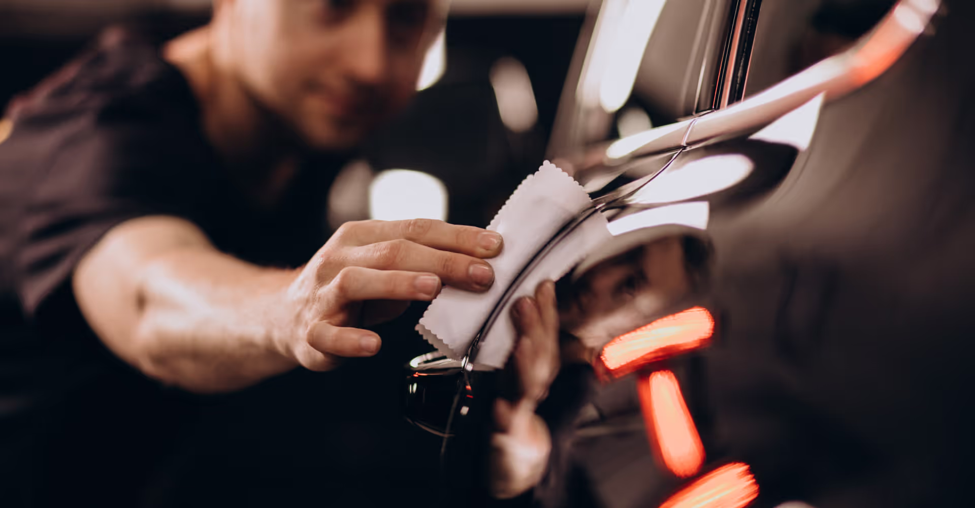Osoba polerująca wygląd czarnego samochodu białą szmatką, odbijającą twarz i czerwone tylne światła.