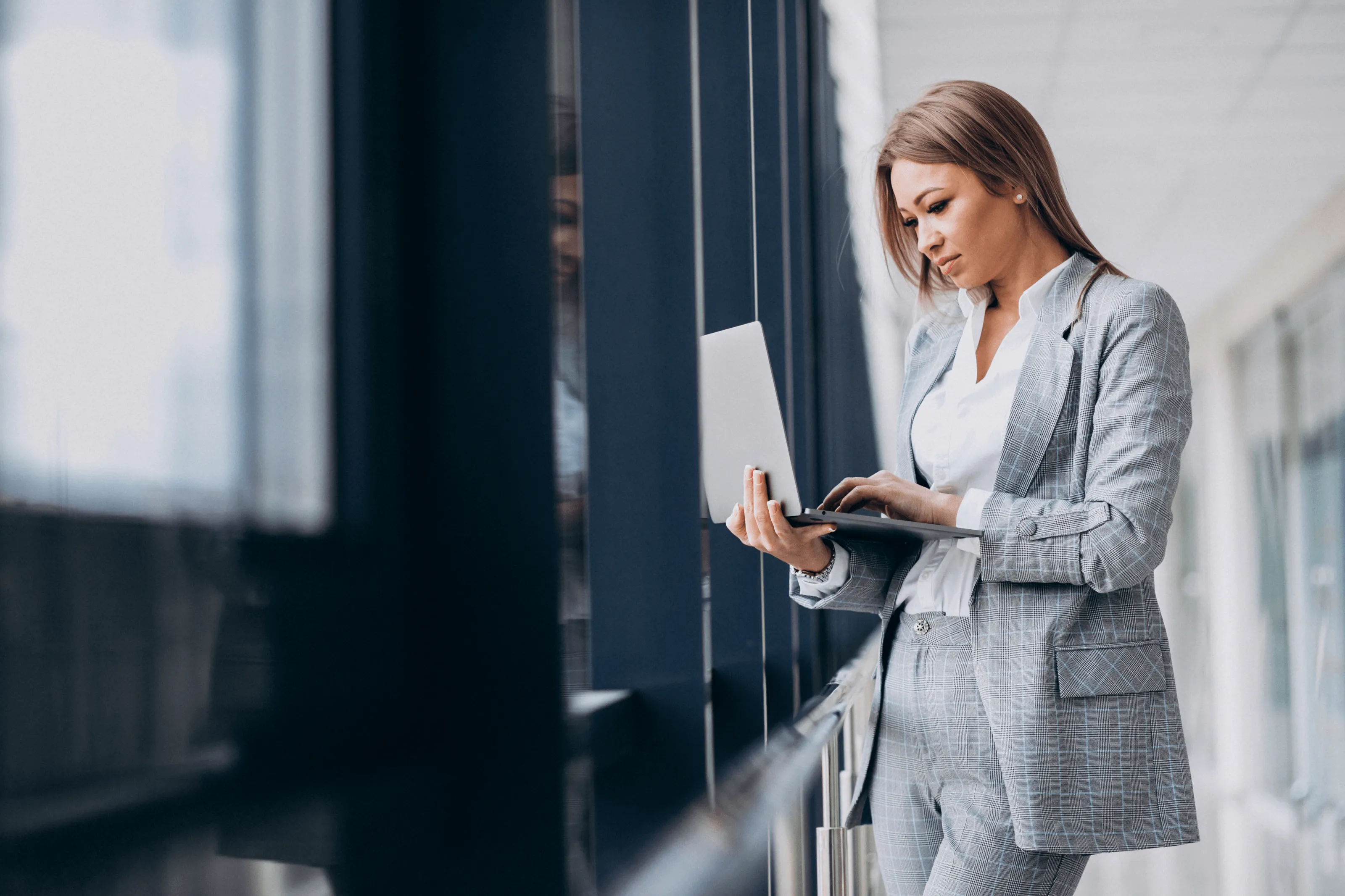 Kobieta w szarym garniturze w kratę stojąca przy oknie podczas pracy na laptopie.
