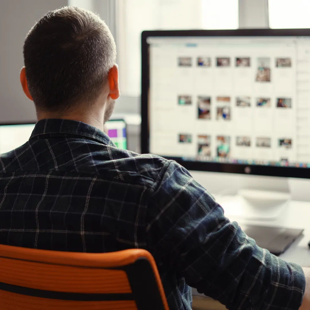 Man in a plaid shirt sitting on an orange chair, working on a desktop computer with multiple image thumbnails on the screen.