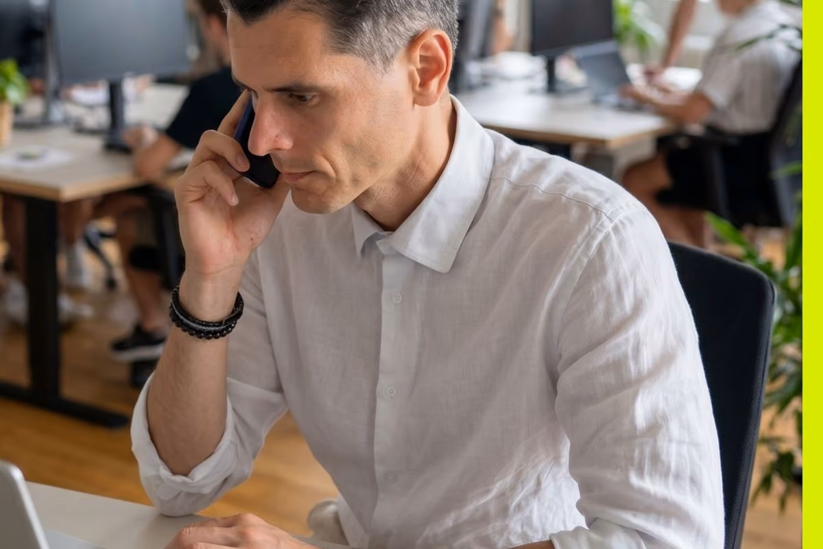 Man in white shirt talking on phone while working on laptop in an office setting.