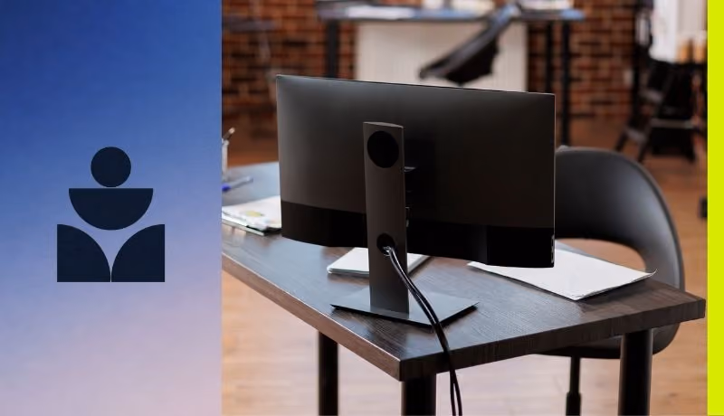 Rear view of a black computer monitor on a dark wooden desk with papers and a chair in background.