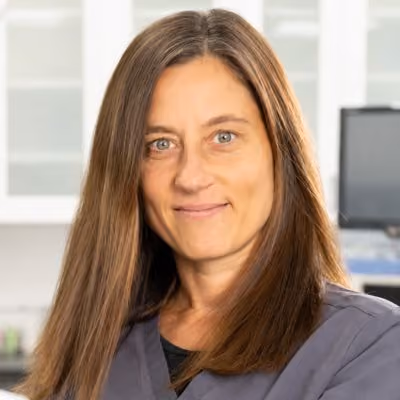 A woman with long brown hair and blue eyes, wearing a dark gray top, smiling at the camera.