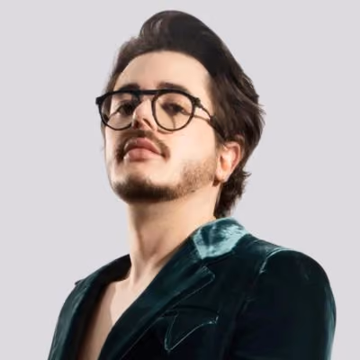 Young man with glasses and facial hair wearing a dark green velvet blazer against a gray background.