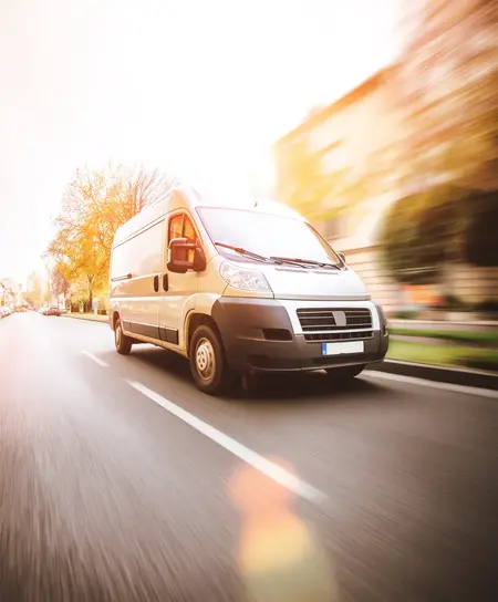 Fourgon blanc roulant rapidement sur une route urbaine bordée d'arbres aux feuilles jaunes en automne.