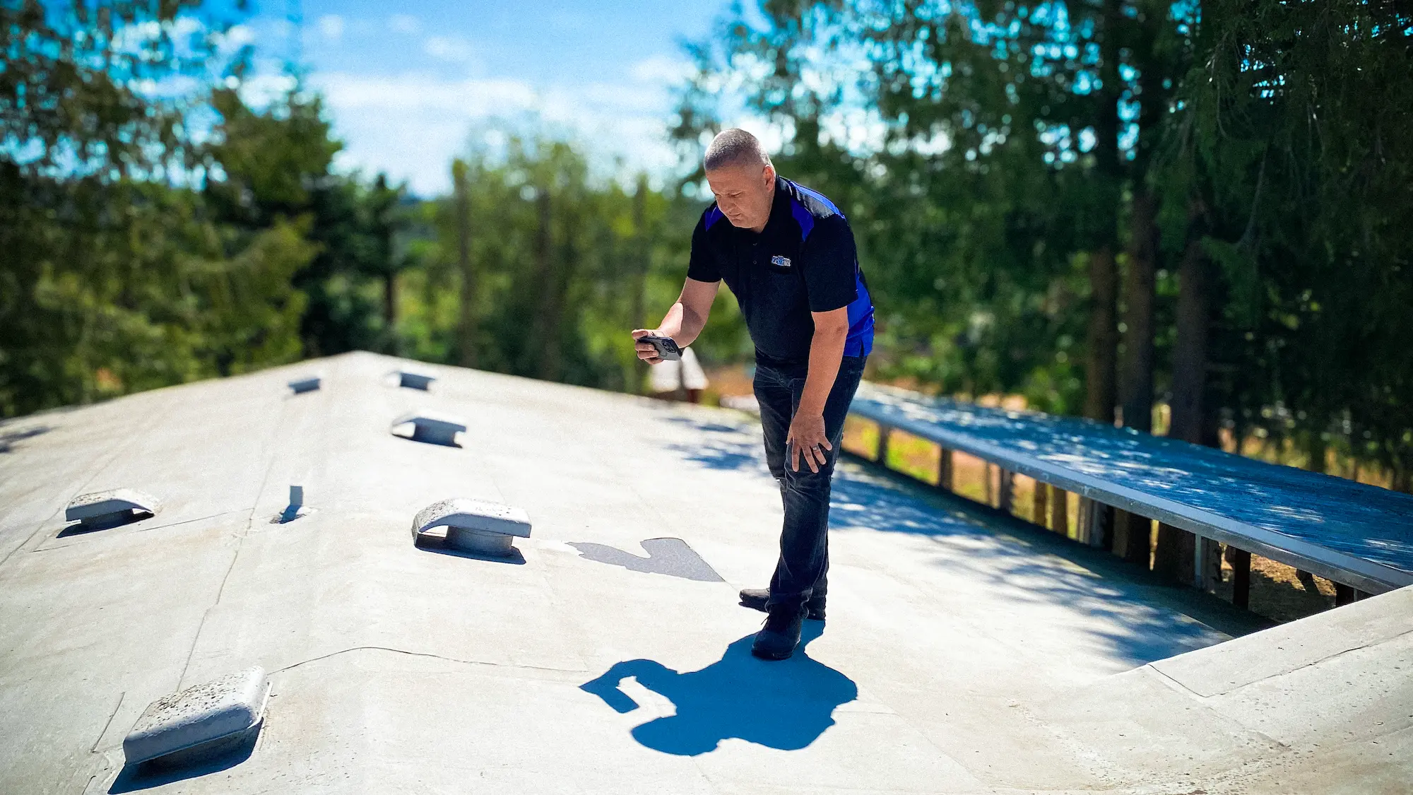 Man wearing gloves and sunglasses power washing a shingles roof under clear blue sky.