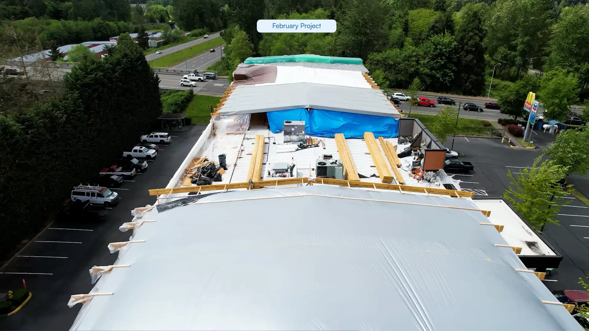 Aerial view of a winter roof under construction with wooden beams, construction materials, and tarps, surrounded by trees and a parking lot.