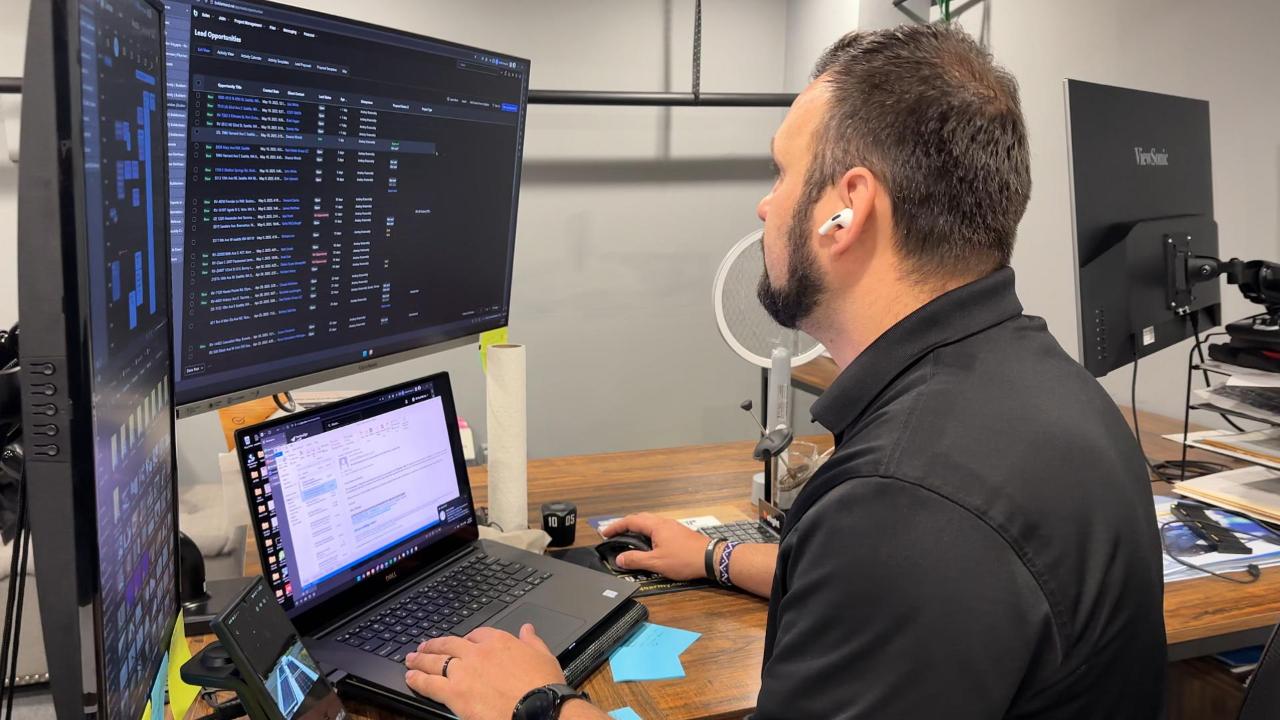 A man, an employee of Flat Roof Pros wearing headphones, works at a desk with two monitors and a laptop, the screens displaying data and emails.