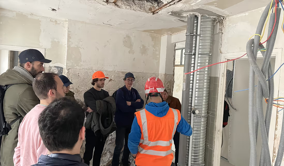 Un groupe de personnes assiste à une visite de chantier dans un bâtiment en rénovation, guidé par un ouvrier portant un casque et un gilet de sécurité.
