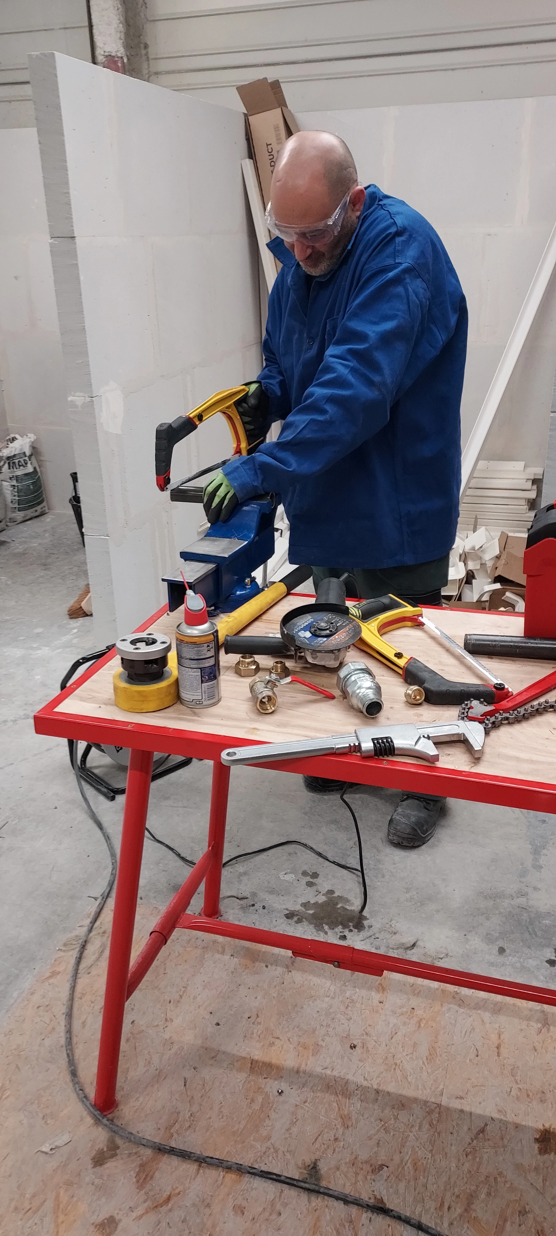 Apprenant en formation professionnelle effectuant une découpe sur établi, équipé d’outils de plomberie et d’équipements de protection.