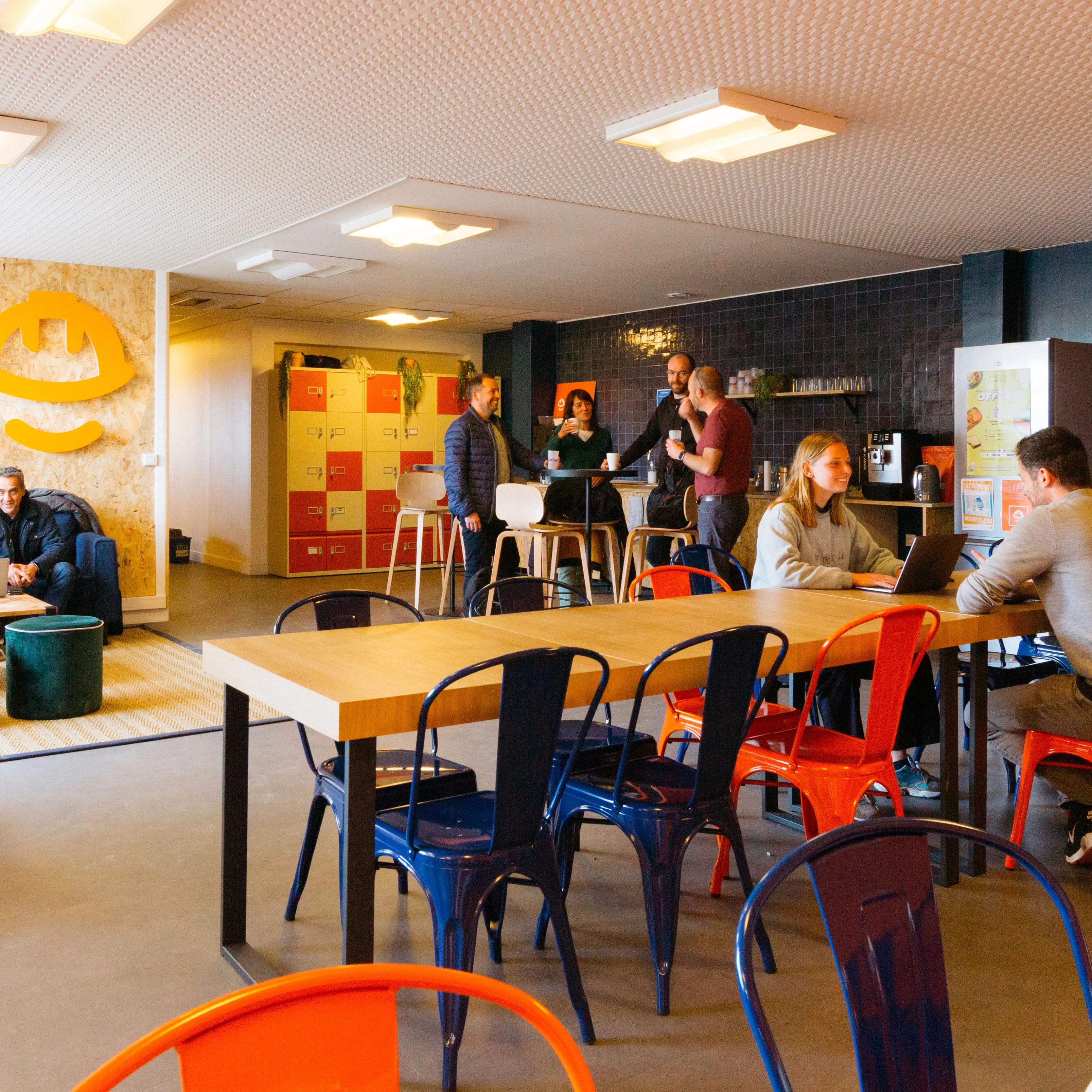 Un espace de coworking moderne et détendu avec des personnes discutant, travaillant sur des ordinateurs portables et prenant un café, entourées de mobilier coloré et d’une décoration chaleureuse.