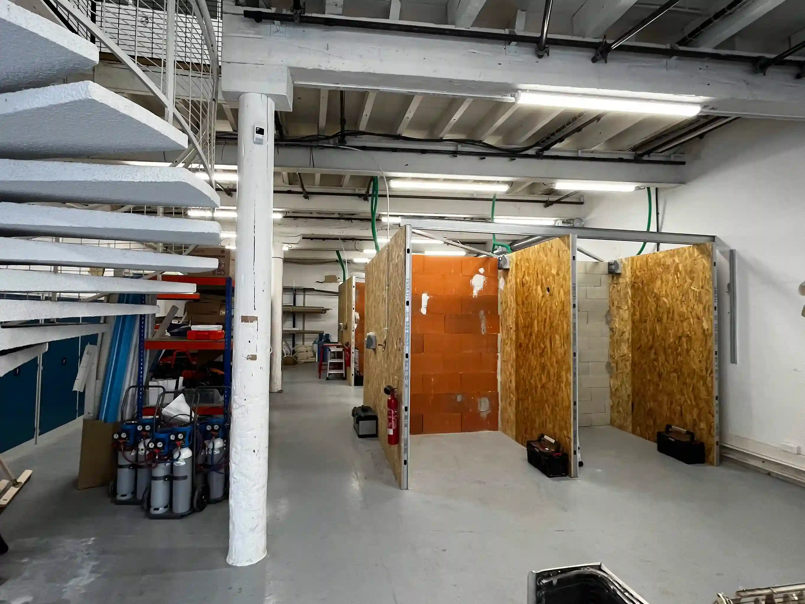Vue d'un plateau technique de formation plomberie-chauffage avec des cabines de pratique en bois et briques, équipées pour l'installation de pompes à chaleur, entourées de matériel professionnel (bouteilles de fluide frigorigène, outillage).