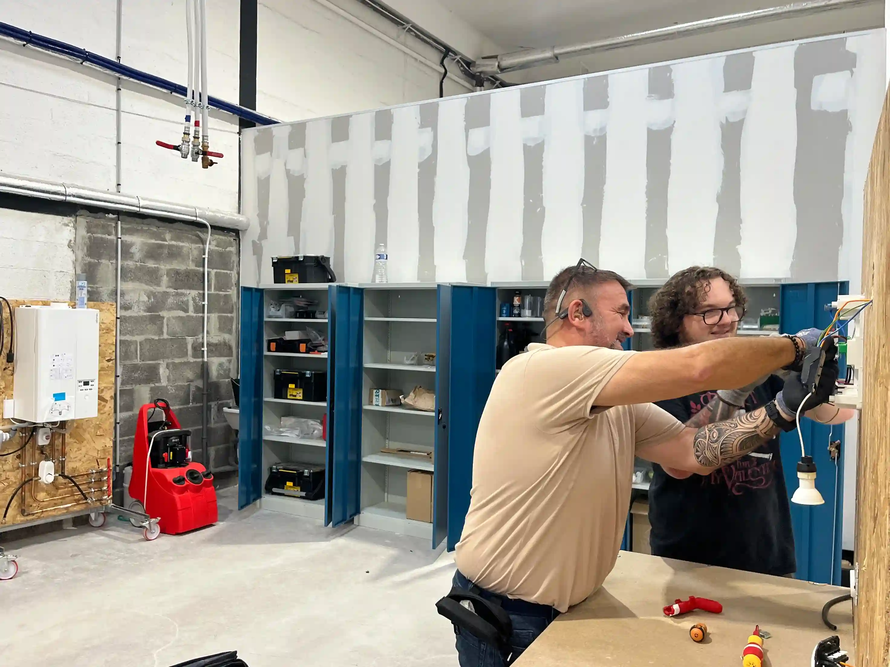 Vue d'une salle de formation en CVC, avec un formateur et un étudiant en train de câbler et de raccorder un tableau électrique, dans un atelier équipé d'une chaudière murale et de tuyauteries cuivre pour la pratique du chauffage et de la plomberie.
