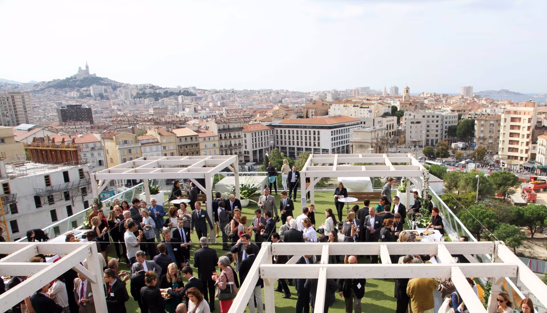 Vue d’un événement professionnel en plein air sur un rooftop à Marseille, avec de nombreuses personnes en tenue de ville échangeant sous des pergolas blanches. En arrière-plan, une vue panoramique de la ville, avec la basilique Notre-Dame de la Garde visible sur la colline.