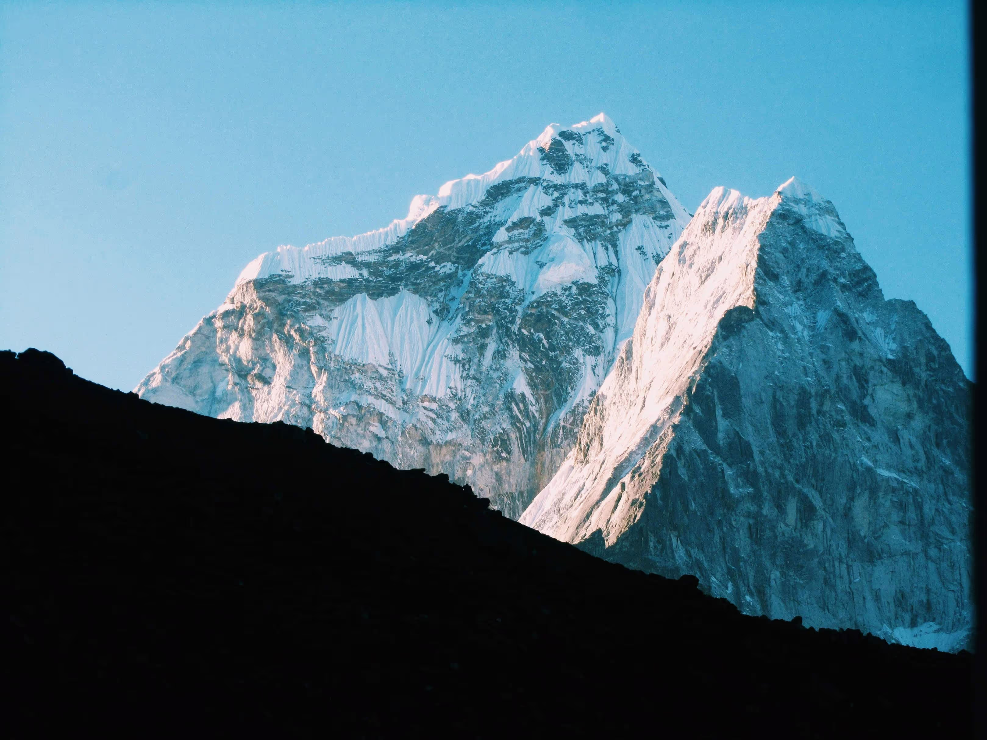Snow-covered mountain peaks illuminated by soft sunlight against a clear blue sky.