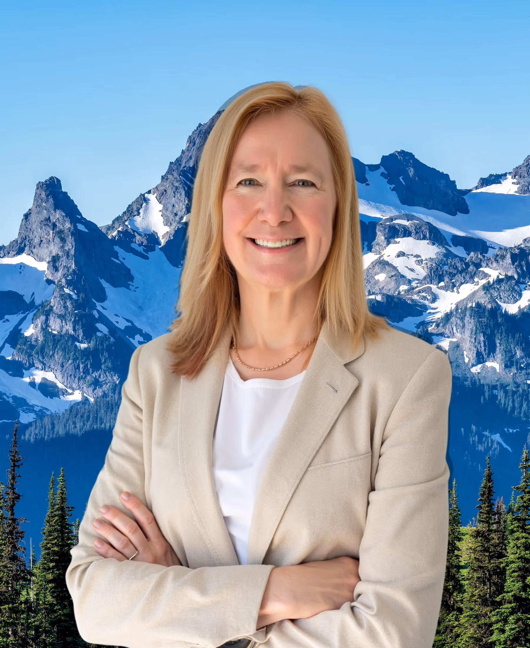 Smiling woman with blonde hair wearing a beige blazer standing with arms crossed in front of snow-capped mountains and evergreen trees.