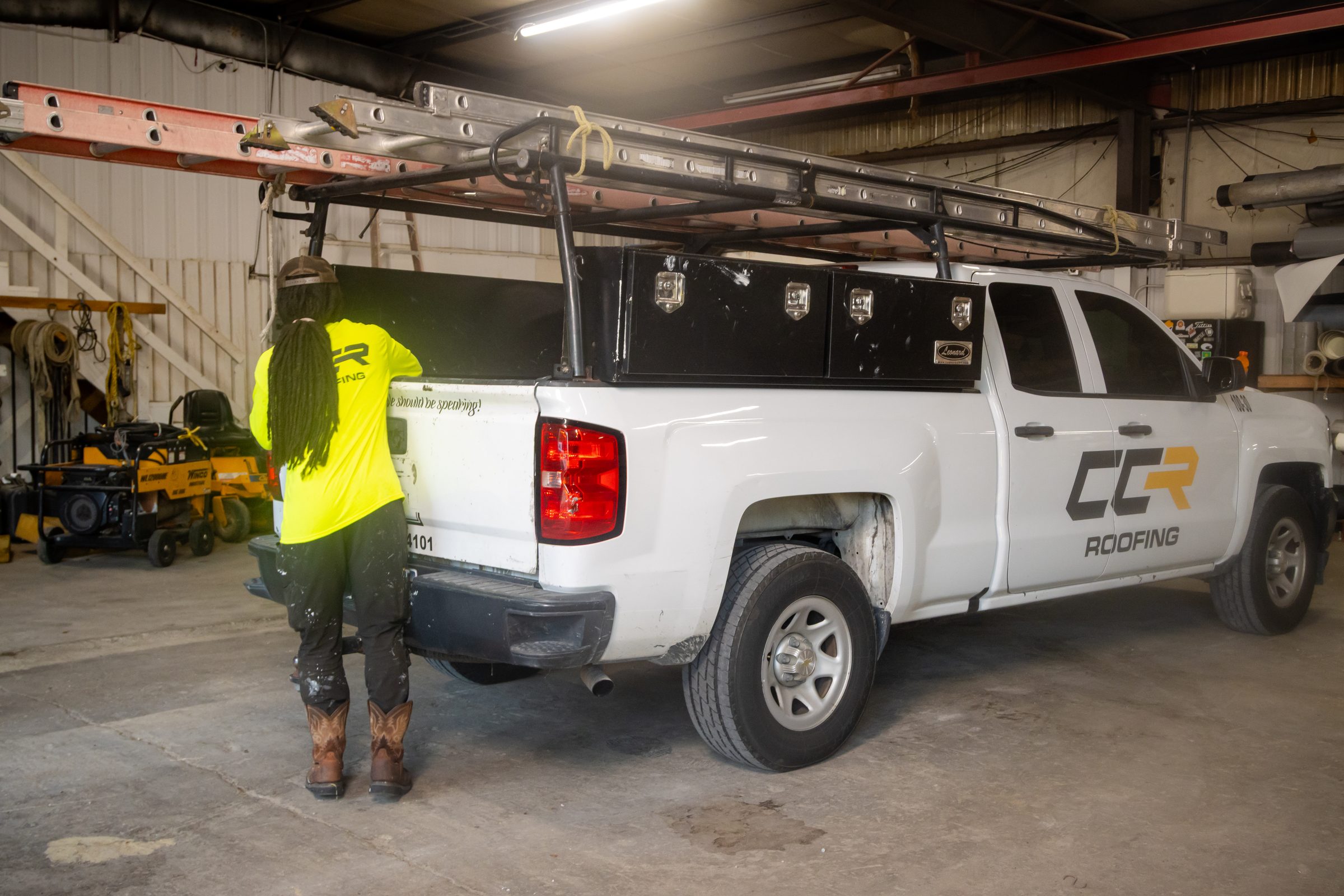 A Coastal Commercial Roofing worker in a high-visibility shirt at the tailgate of a CCR Roofing pickup truck loaded with ladders, inside the company warehouse.