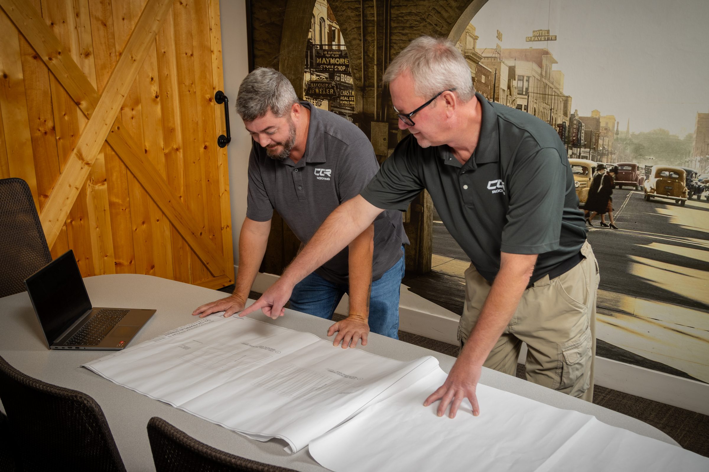 Two Coastal Commercial Roofing team members in branded polo shirts reviewing blueprints spread across a table in the company office.