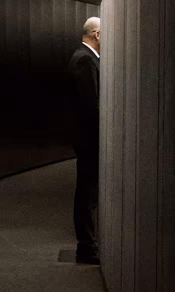 Homme en costume noir debout dans un coin sombre, partiellement caché derrière un panneau gris.