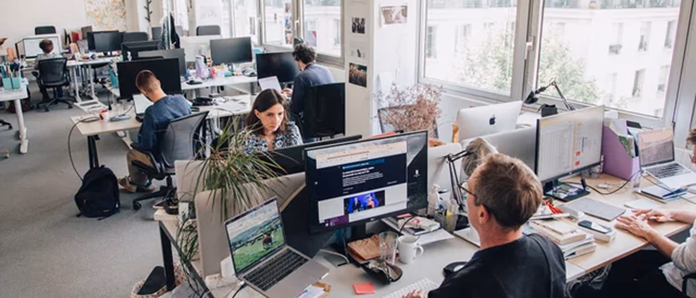Bureau moderne avec plusieurs personnes travaillant sur des ordinateurs et des laptops près de grandes fenêtres lumineuses.