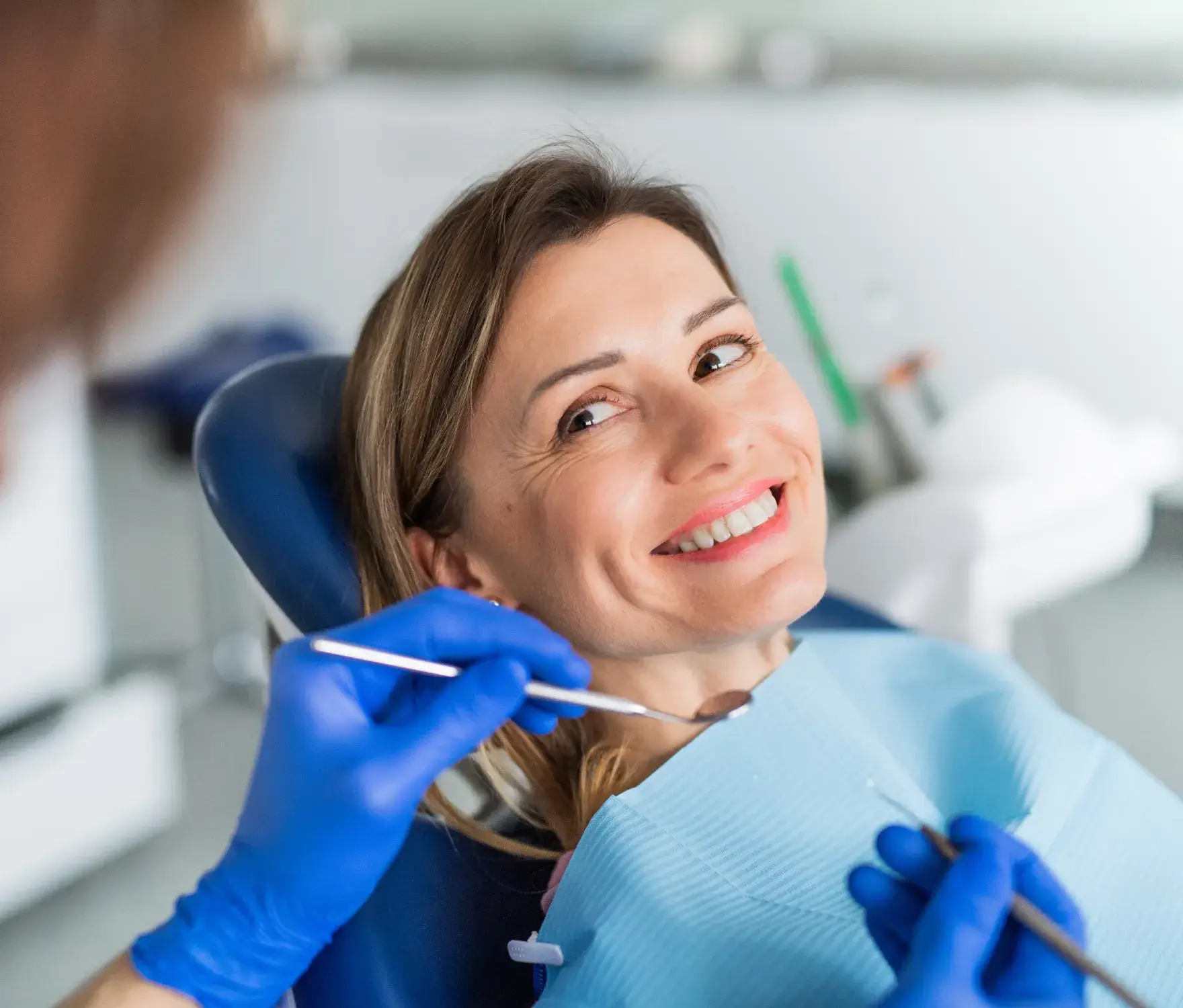 A dentist examines a smiling woman’s teeth while she sits in a dental chair.
