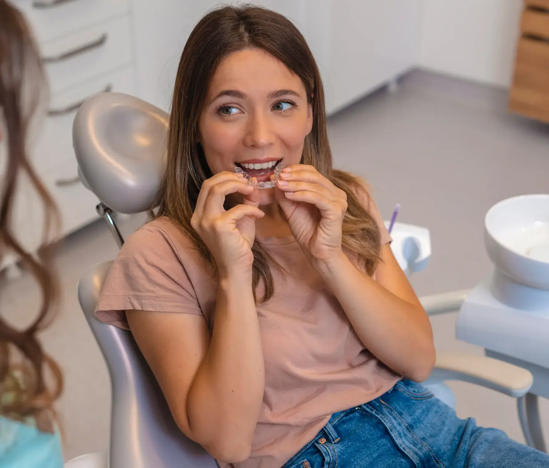 A woman sitting in a dental chair, holding a clear dental aligner near her mouth.