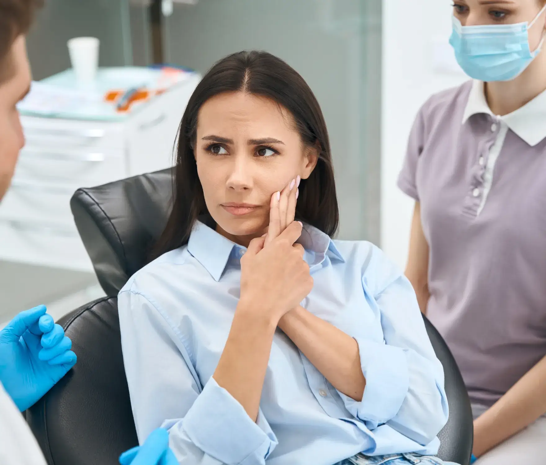 A woman holding her jaw looks concerned while sitting in a dental chair with two dentists present.