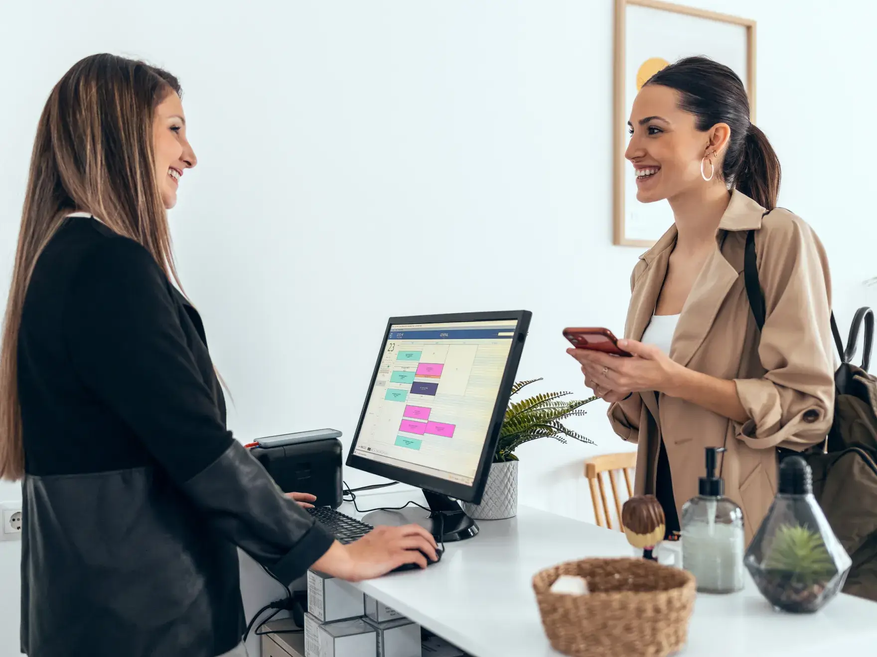 Two women smiling and talking at a reception desk, one using a computer and the other holding a phone.