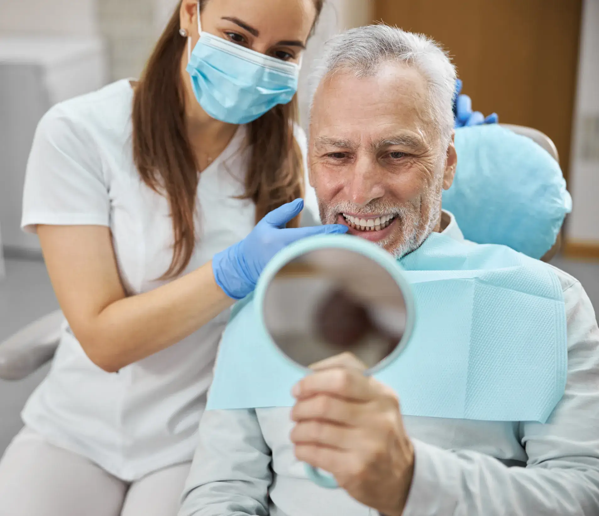 A dentist helps an older man examine his teeth in a handheld mirror.