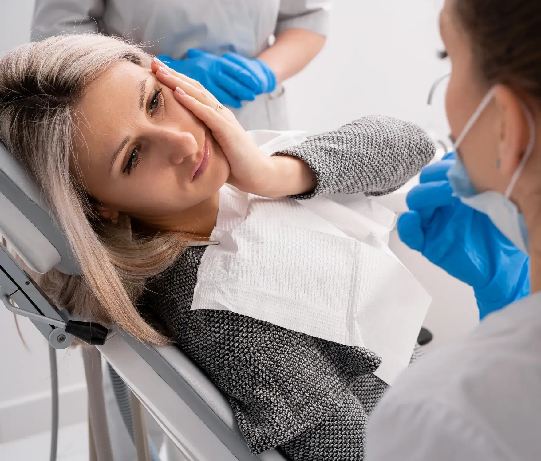 Woman sitting in a dental chair, holding her cheek, while talking to a dentist wearing gloves.