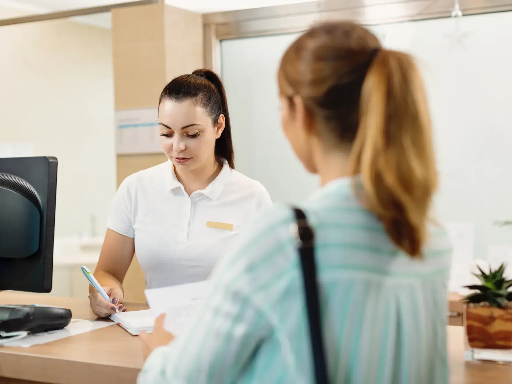 A receptionist in a white shirt is writing as she assists a customer at the desk.