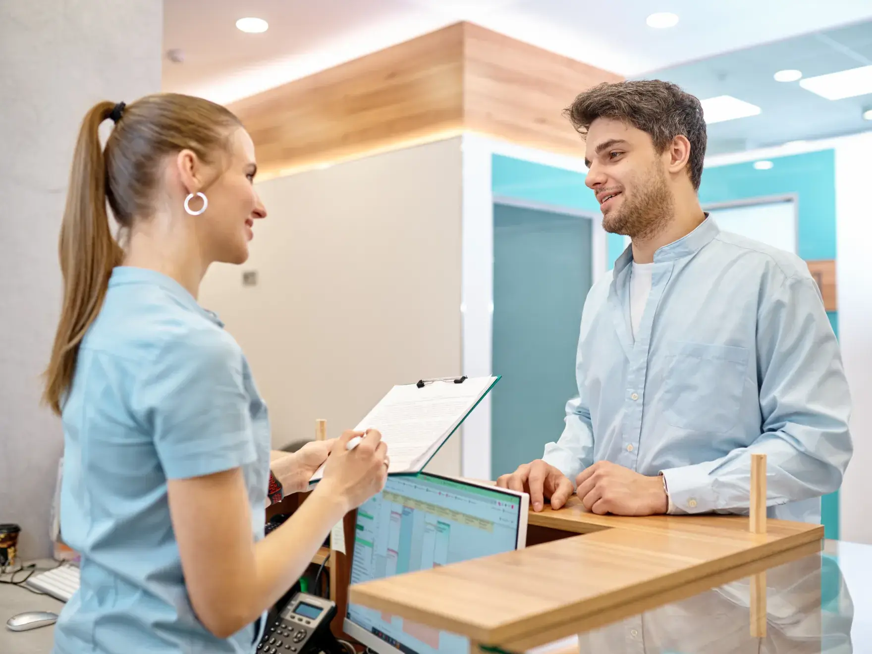 A woman at a reception desk talks to a man, holding a clipboard with paperwork.