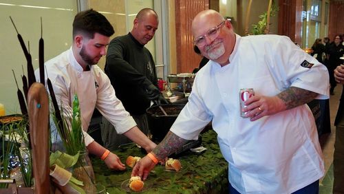 A chef smiling as two others prepare food in the background at an event with the American Culinary Federation New Orleans Chapter