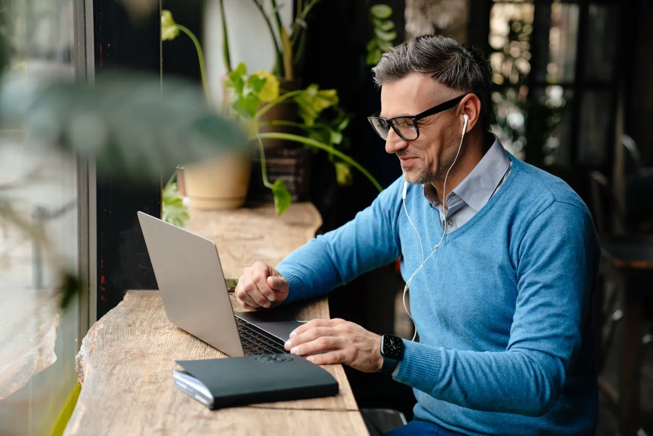 Man wearing glasses and blue sweater using a laptop at a wooden counter with a notebook nearby.
