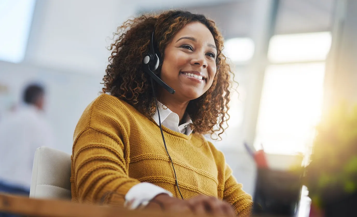 Smiling woman wearing a headset and mustard sweater working in a bright office.