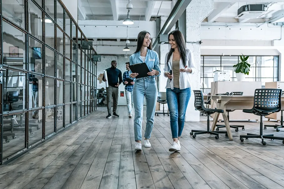 Two young women walking and talking in a modern office space with wooden floors and glass-walled rooms.