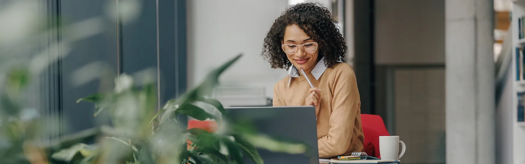Smiling woman with glasses working on a laptop at a desk with a plant in the foreground.