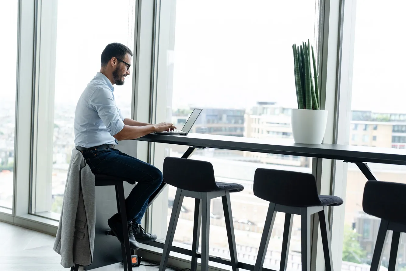 Man wearing glasses and a light blue shirt sitting on a high stool by a tall window using a laptop at a long counter with a potted plant.