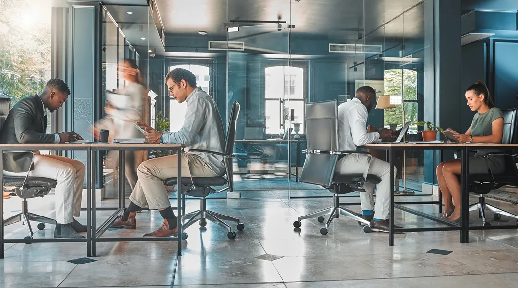 Four professionals seated at desks in a modern office with glass walls, two working on laptops and two using tablets, while a blurred figure walks by.