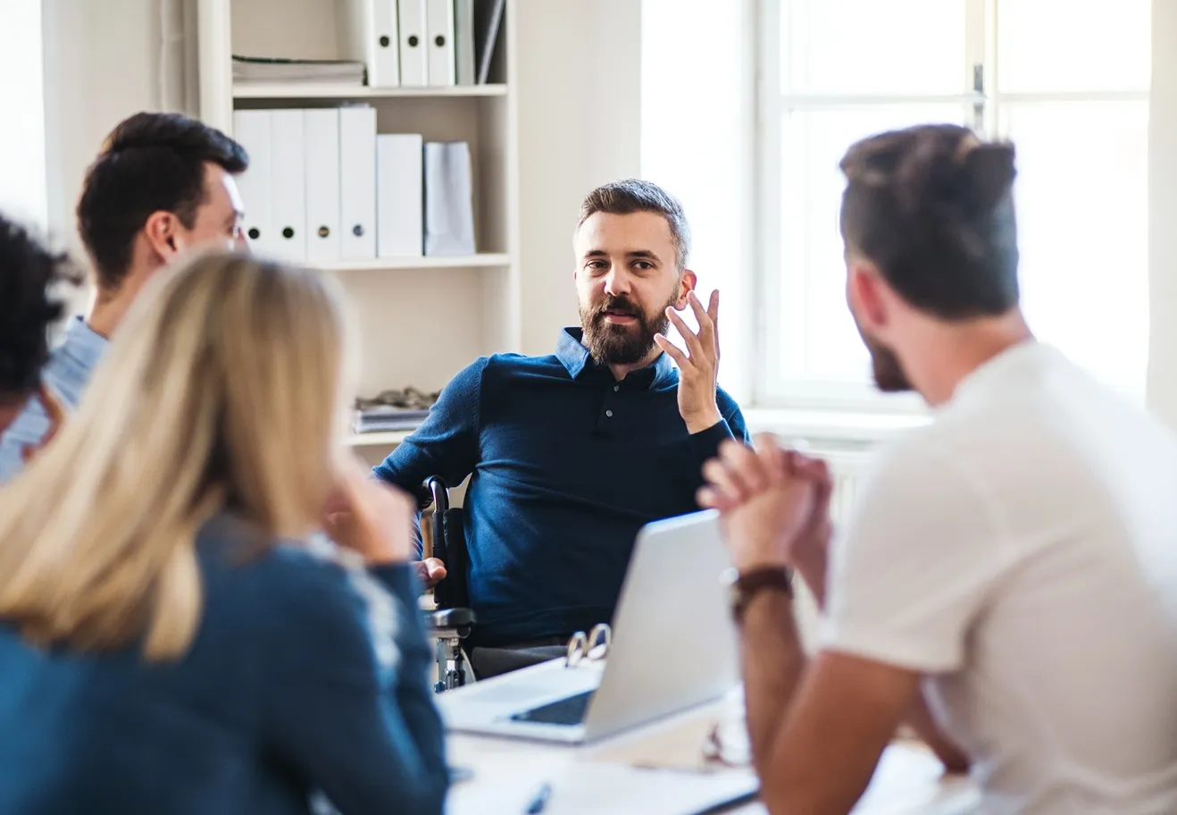 Man in wheelchair leading a discussion with colleagues around a table in a bright office.