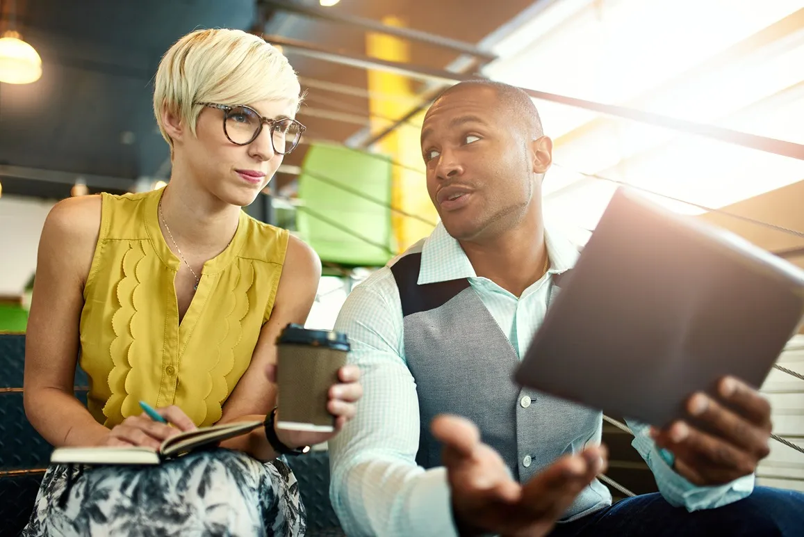 Man and woman in an office space working together on a tablet and writing notes