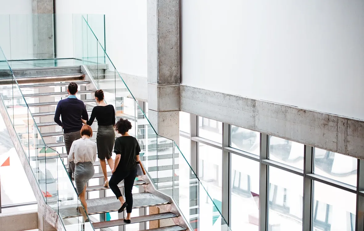 Four professionally dressed adults ascending a modern glass and metal staircase in a bright office building.