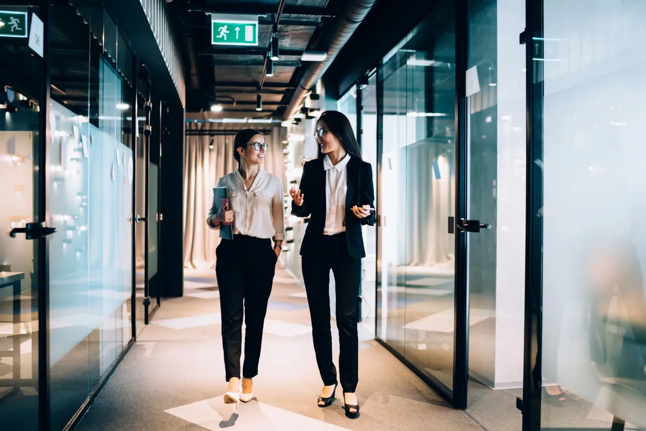 Two women walking down the hall of an office carrying notebooks and laptops