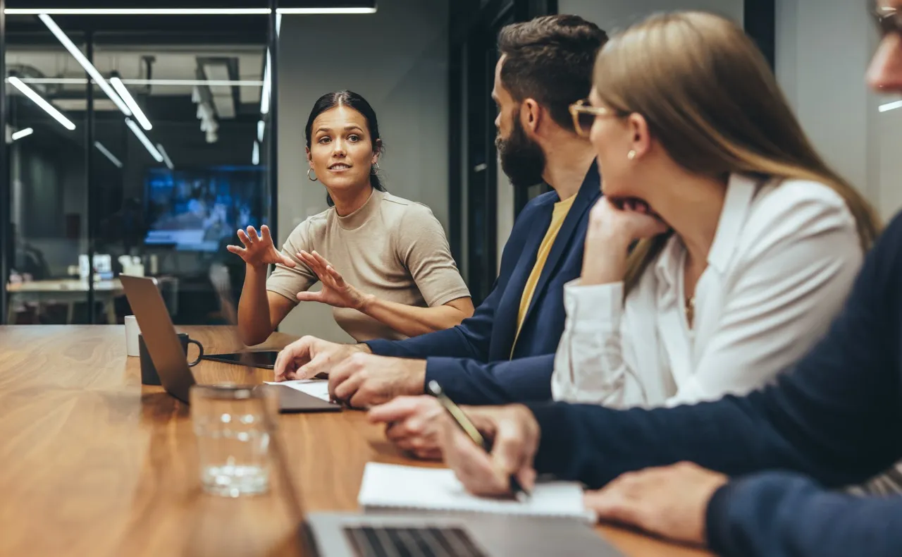 Multiple people in meeting room sitting at the table looking a woman speaking