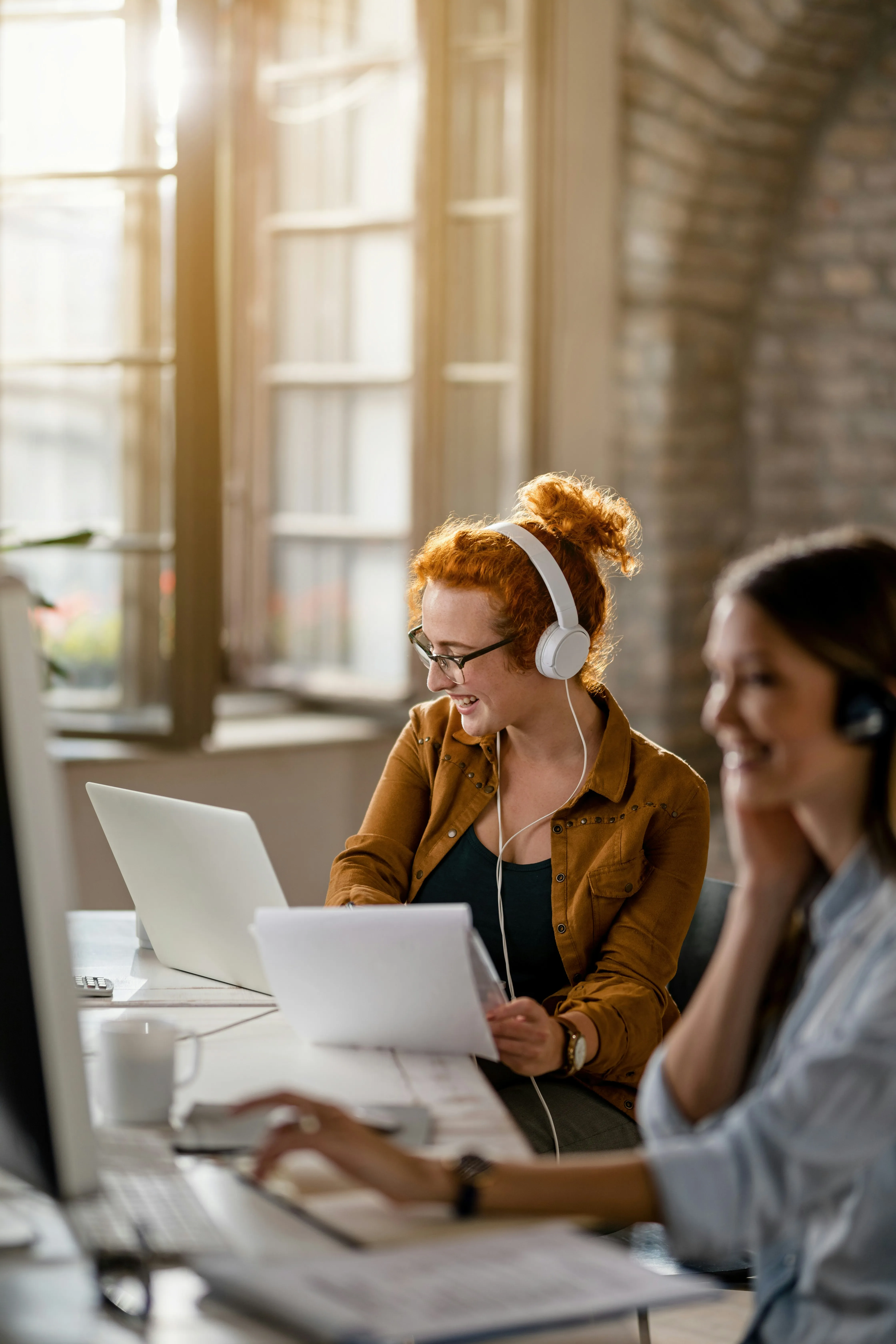 Two women working at computers in a bright office, one with red hair wearing headphones and holding papers, the other blurred in the foreground.